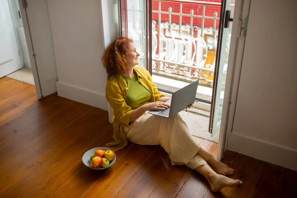 Senior Woman Relaxing With Laptop And Fruits On Floor Near Open Window At Home, Dreamy Smiling Elderly Female Using Computer For Leisure, Enjoying Domestic Pastime And Retirement, Free Space