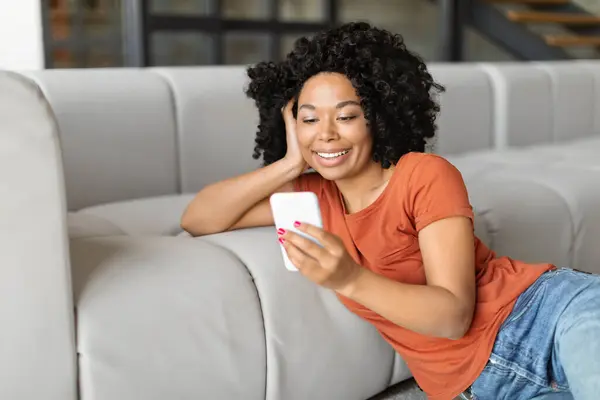 Smiling Black Woman Watching Videos On Smartphone At Home, Happy African American Female Sitting On Floor And Leaning At Couch, Enjoying Online Content While Relaxing In Living Room, Closeup