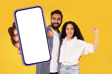 Neşeli genç Arap eşler ellerinde boş dokunmatik ekranlı büyük bir cep telefonu tutuyorlar, modern alet kavramlarını gösteriyorlar ve işaret ediyorlar, evet, sarı arka planda stüdyo çekimi, kolaj