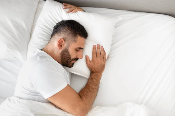 Bearded man sleeps in his bed, hugging a pillow for comfort in bedroom indoors, resting covered with soft blanket. Concept of healthy restful sleep and wellbeing. View from above