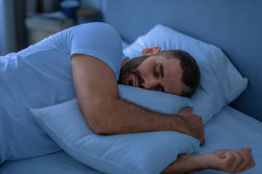 Huzur içinde uyuyan, yastığı kucaklayan, geceleri yatakta yatan adam. Dinlenme rutini, uyku ve dinlenme. Milenyum adamı rahat, modern yatak odasında huzur içinde dinleniyor. Düşük Işık