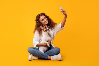 Neşeli, kıvırcık saçlı kadın, neşeli Jack Russell Terrier 'i ile bacakları çapraz oturarak, neşeli sarı arkaplan karşısında selfie çekerek, eğlenceli bir anı yakalıyor.
