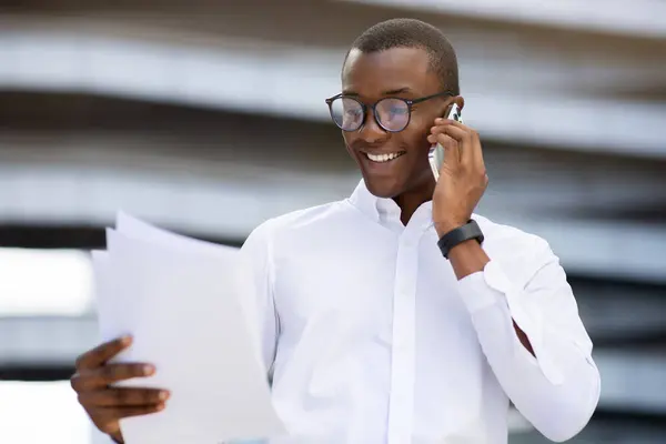 Business Lifestyle. Handsome black businessman talking on cellphone and reading documents outdoors, smiling african american male entrepreneur having phone call with business partner, copy space