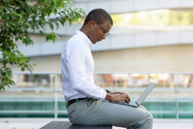 Modern ofis geçmişiyle dışarıda otururken, dizüstü bilgisayarı üzerinde çalışan konsantre siyah işadamı Afro-Amerikan erkek girişimci uzaktan kumanda için bilgisayar kullanıyor.