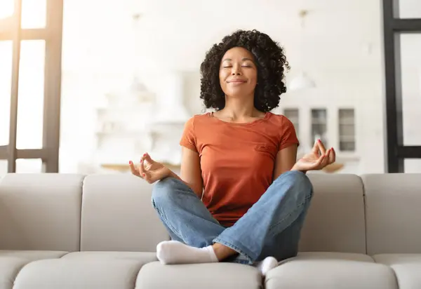 Stress Relief Concept. Peaceful young black woman in casual clothes sitting cross-legged on couch, smiling calm african american female practicing meditation with closed eyes, enjoying home comfort