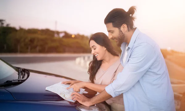 Smiling middle eastern couple standing by their car at dusk, joyfully planning their road trip with a map spread on the hood, panorama