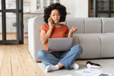 Duygusal siyah kadın dizüstü bilgisayarla sorun yaşıyor, oturma odasında oturma odasında otururken müşteri desteğini arıyor, endişeli Afrikalı Amerikalı kadın bilgisayar kullanıyor ve cep telefonuyla konuşuyor.