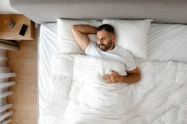 Huzurlu orta yaşlı bir adamın modern yatak odasında şekerleme yapması. Yatakta rahat bir şekilde uzanmış kafasının arkasında el ele tutuşmuş, rahat çarşaflara sarılmış. Dinlenme ve rahatlama, sağlıklı uyku.