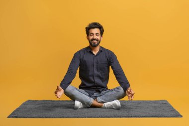 Rahat genç Hintli adam tek başına meditasyon yapıyor, halı üzerinde bacak bacak üstüne atmış oturuyor, yakışıklı doğulu adam yoga yapıyor, sarı stüdyo arka planında izole edilmiş poz veriyor, uzay kopyaları yapıyor.