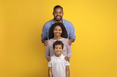 Mutlu siyah aile, anne baba ve oğul sırada beklerken kucaklaşıyorlar, neşeli Afrikalı Amerikalı ebeveynler ve ergen erkek çocuk birbirlerine bağlanıyorlar, sarı stüdyoda poz veriyorlar, boşluğu kopyalıyorlar.