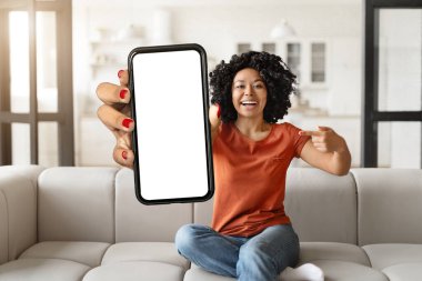 Neşeli genç siyah kadın, evde koltukta otururken büyük beyaz ekranlı akıllı telefonu işaret ediyor, gülümseyen Afrikalı Amerikalı kadın, yeni mobil uygulama, kolaj ve model için kopyalama alanı gösteriyor.