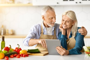 Neşeli yaşlı çift mutfakta sağlıklı öğle yemeği pişirirken dijital tablet kullanıyor, internette yemek arıyor, kıdemli eşler birlikte yemek hazırlıyor ve evde eğleniyor,