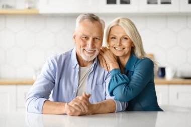 Mutlu kıdemli eşlerin mutfak masasında poz verişi, neşeli yaşlı bir çiftin kameraya sarılıp gülümsemesi, evde birlikte vakit geçirmesi, emekliliğin tadını çıkarması, yakın plan çekimleri...