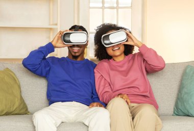 Afro-Amerikalı karı-koca sanal gerçekliği deneyimliyor, sanal gerçeklik cihazlarını evde oyun oynamak için kullanıyor, kanepede oturuyor ve gülümsüyor. Modern dijital eğlence ve eğlence.