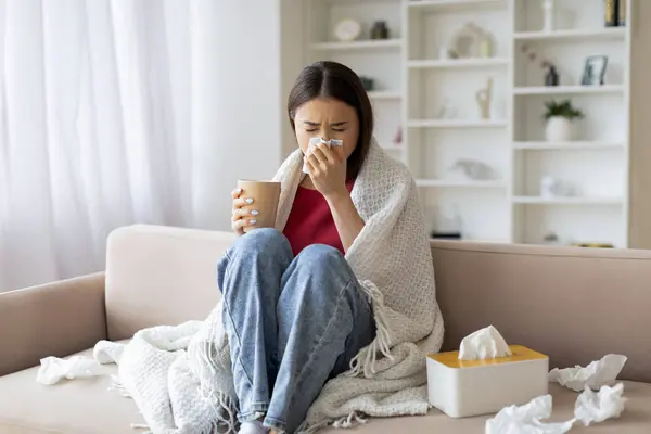 Cold And Flu Concept. Sick asian woman blowing her running nose in paper tissue and drinking tea at home, ill korean lady covered in blanket sitting on couch in living room, feeling unwell