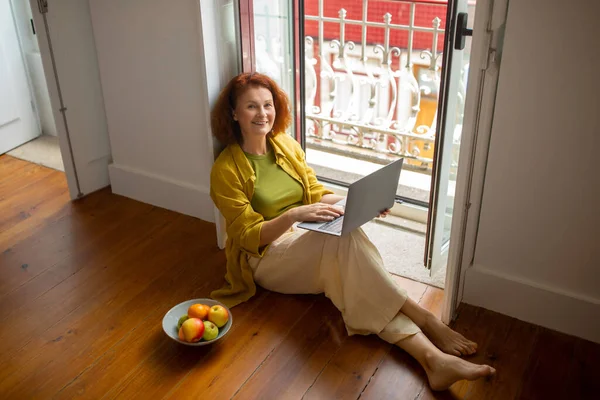 Happy senior lady with laptop computer sitting on floor by opened window at home, smiling older woman working online or browsing internet while relaxing in minimalistic interior, above view