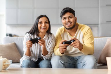 Neşeli Hintli çift evlerinde joysticklerle video oyunu oynuyor, oturma odasında otururken mutlu doğu erkeği ve kadını eğlendiriyorlar, neşeli eşler oyun oynuyorlar, yakın çekim yapıyorlar.