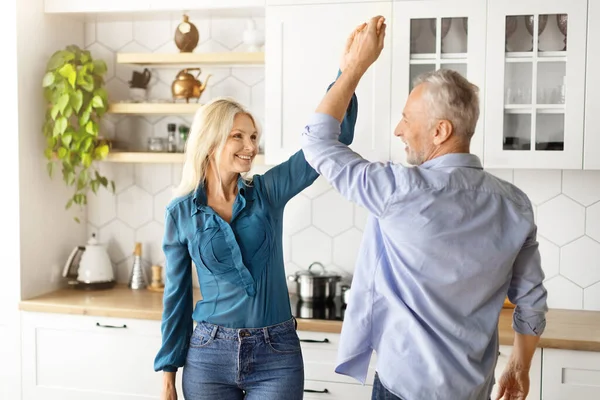 Portrait Of Cheerful Older Couple Dancing Together In Kitchen Interior, Happy Romantic Senior Spouses Having Fun, Holding Hands And Smiling To Each Other, Enjoying Spending Time At Home