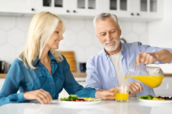 Cheerful Older Spouses Enjoying Healthy Breakfast Together In Kitchen, Happy Senior Couple Enjoying Delicious Food At Home, Caring Husband Pouring Orange Juice From Jug, Closeup Shot