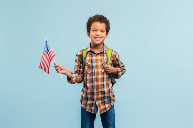 Amerikan bayrağı tutan neşeli genç bir çocuk, ekose gömlek ve sırt çantası giyiyor, gururlu ve mutlu görünüyor, yumuşak mavi arka planı vatanseverliği ve yabancı dilleri simgeliyor.