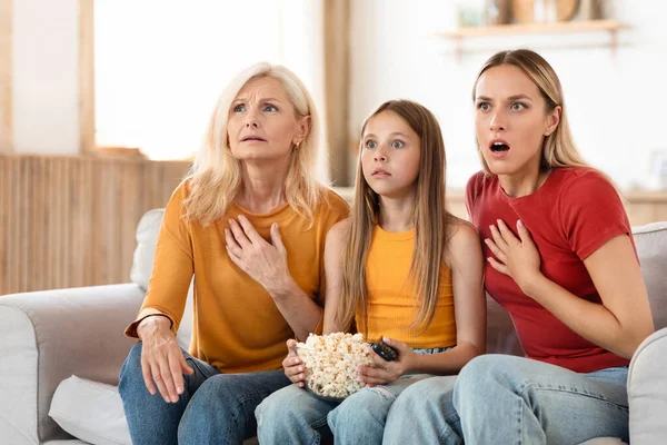 Shocked family three generations of women watching TV together at home, grandmother, mother and grandchild teen girl sitting on couch, eating popcorn, watching thriller