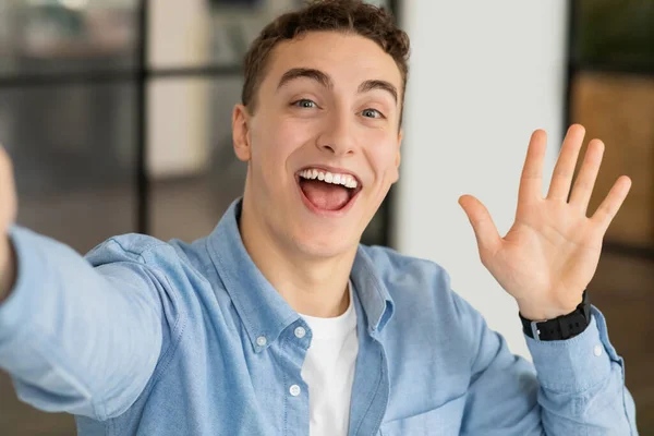 Smiling attractive millennial caucasian guy manager make photo at smartphone, waving hand in modern coworking office. Work with gadget, business app, say hello and video call