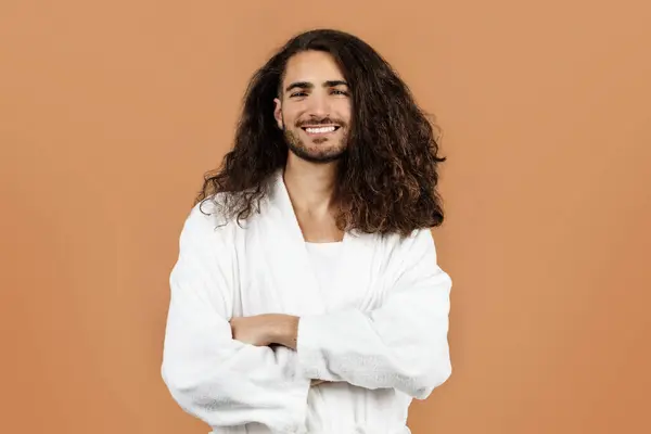 Studio portrait of cheerful bearded young guy with curly long brunette hair, poses in white bathrobe after shower and morning beauty routine against beige backdrop. Spa and pampering