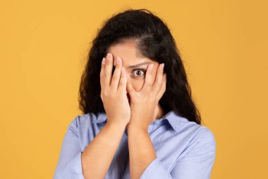 Korkmuş, şok olmuş, bin yıllık Latin esmer kadın elleri kapalı, tek gözü kapalı, turuncu stüdyo arka planında izole edilmiş, yakın plan. İnsanlardan korkmak duygu, kötü haber