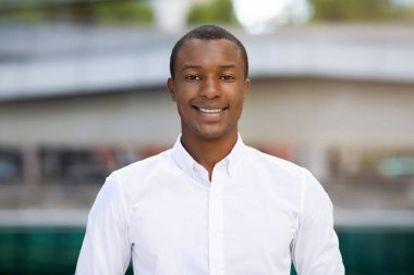 Beyaz tişörtlü mutlu genç adamın portresi, Milenyum Afrikalı Amerikalı iş adamı kameraya bakıyor ve gülümsüyor, ofisin arka planında ayakta duruyor.