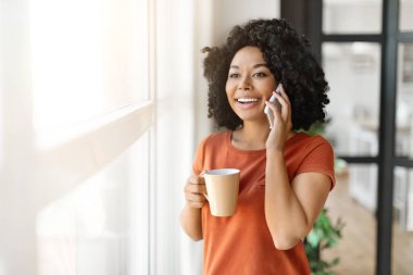 Güzel, genç, siyah bir kadın kahve içiyor ve evinin penceresinin yanında cep telefonuyla konuşuyor gülümsüyor, Afro-Amerikan bir bayanın keyifli mobil sohbetleri ve sıcak içeceklerin tadını çıkarıyor,