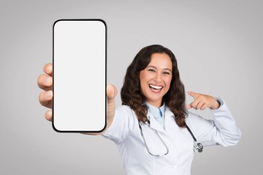 Heyecanlı kadın doktor gri arka planda dururken kamerada boş ekranla akıllı telefonu gösteriyor. Kadın doktor telefonu işaret ediyor ve uygulama, model öneriyor.