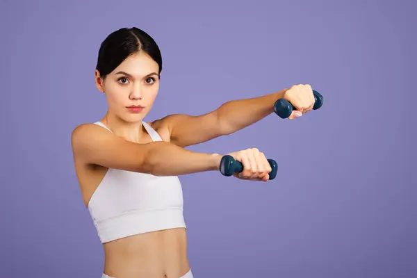 严重的高加索瘦女人在运动服做运动与手哑铃 看着相机孤立在紫色工作室的背景 肌肉运动 体重减轻和身体护理 — 图库照片