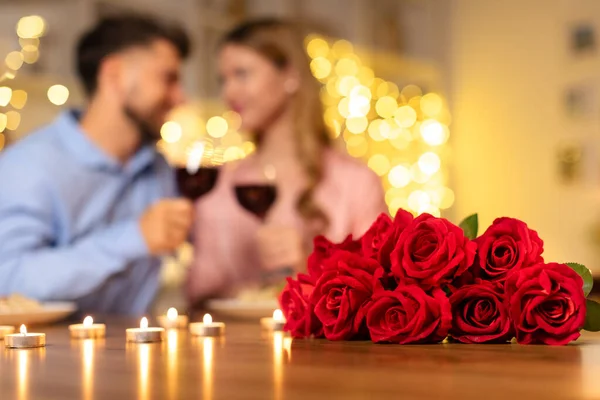 Blurry couple toasting wine glasses behind vivid bouquet of red roses, surrounded by candles, celebrating romantic evening at home