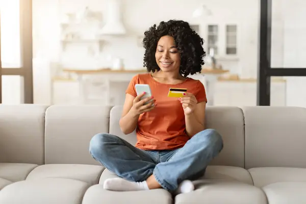 Mobile Payments. Smiling Black Woman With Smartphone And Credit Card In Hands Sitting On Couch At Home, Happy Young African American Female Making Internet Shopping, Enjoying E-Commerce, Copy Space