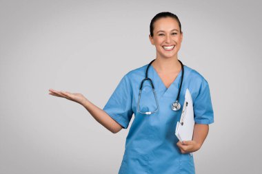 Heyecanlı Avrupalı kadın doktor elinde bir not panosuyla boş avuçlarda, boş alanlarda, reklam ürünlerinde ya da kliniklerde gri stüdyo arka planında izole edilmiş poz veriyor.