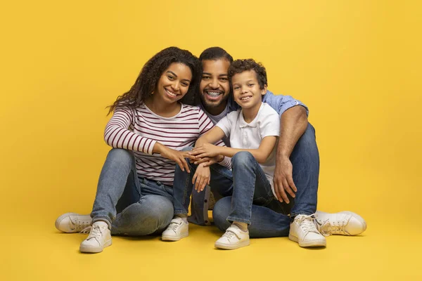 笑顔の黒い親と小さな息子の肖像画 黄色い背景の上に一緒にポーズ スタジオで床に座っている間 3つのボンディングの幸せな若いアフリカ系アメリカ人の家族 閉じるショット — ストック写真