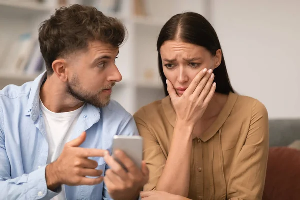 Jealous young husband showing cheating messages on phone to wife indoor, suspecting affair and demanding to explain messages. Man struggling with trust in relationship checking womans cellphone