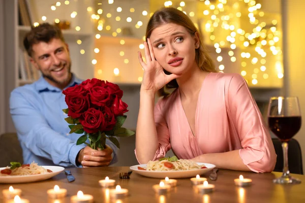 Woman appears unconvinced as smiling man offers bouquet of red roses during candlelit dinner with a hint of romantic tension