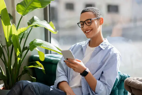 Happy calm millennial latin woman in glasses enjoy peace and spare time, typing on smartphone, chat in social networks in modern eco cafe, office interior. Relax, blog on device