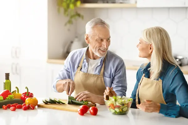 Portrait of happy senior couple cooking food in kitchen at home, happy elderly man and woman wearing aprons preparing lunch or dinner at home, chopping vegetables and smiling to each other