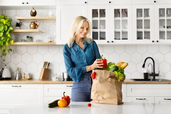 Cheerful senior woman unpacking paper bag full of organic vegetables in kitchen, happy elderly lady checking fresh groceries after food shopping, standing near table at home, copy space