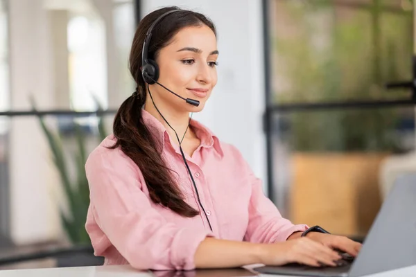 Smiling millennial arab woman manager in headset typing on laptop, enjoy work in modern coworking office. Business support, job service remotely, lifestyle with device, career