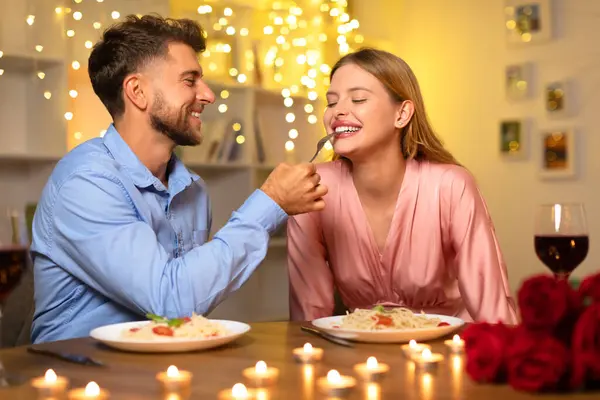 Laughing man tenderly offers fork of spaghetti to his partner during a candlelit dinner, surrounded by a bokeh of twinkling lights on background