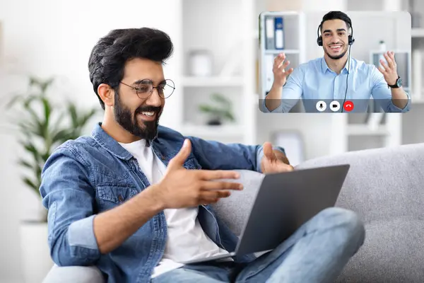 Cheerful Indian Guy Have Video Call With Tech Support. Using Laptop, Sitting On Sofa At Home. Student Learning Online Using Computer, Calling Teacher Middle Eastern Man
