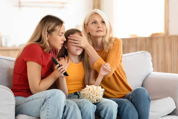 Family three generations of women watching TV together at home, grandmother, mother and grandchild teen girl sitting on couch, eating popcorn, watching horror, adult women covering child eyes