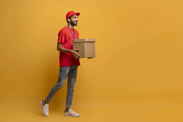 Smiling delivery man in red uniform and cap carrying a cardboard box, happy handsome courier guy with package in hands walking on bright yellow background in studio, advertising delivery services