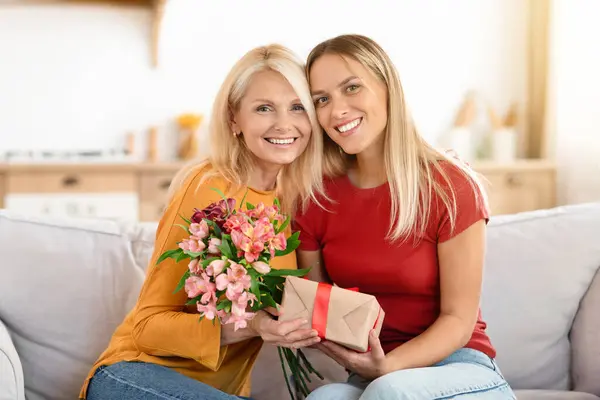Loving young adult woman congratulate excited elderly lady with Mothers Day at home. Smiling caring grownup millennial daughter present gift flowers to old mom on womens day