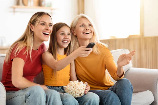 Positive three generations of women family watching TV together at home, happy mother, teen daughter and grandmother sitting on couch with remote control and bowl with popcorn, copy space