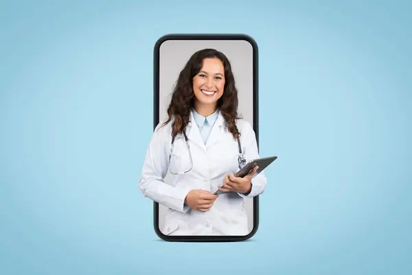 stock image Cheerful european woman doctor in white coat with stethoscope and tablet on big phone screen on blue background. App for medical help, diagnostics, exam, ad and offer