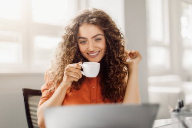 Kahvesini yudumlarken gülümseyen kıvırcık saçlı neşeli genç bir kadın, parlak bir ofis alanında dizüstü bilgisayarının başına oturmuş, hoş bir çalışma ortamını işaret ediyor.